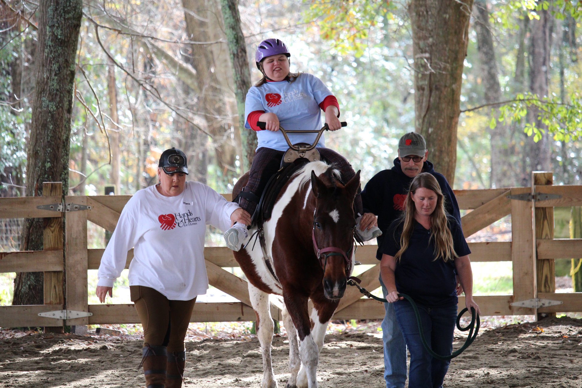 One person sits on a horse with an assistive device and two people walk next to the hoprse 